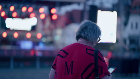 close up of a older caucasian woman with glasses walking towards the stage and back with flickering lights while looking at phone in slow motion during a night in historical city bardejov, slovakia