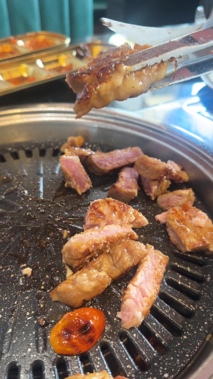 Des pinces et des ciseaux coupent de la poitrine de porc grillée sur un barbecue coréen de table, sous un éclairage intérieur lumineux.