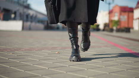 close-up leg view of individual wearing black boots and coat, holding black briefcase, walking along interlocked path in urban environment