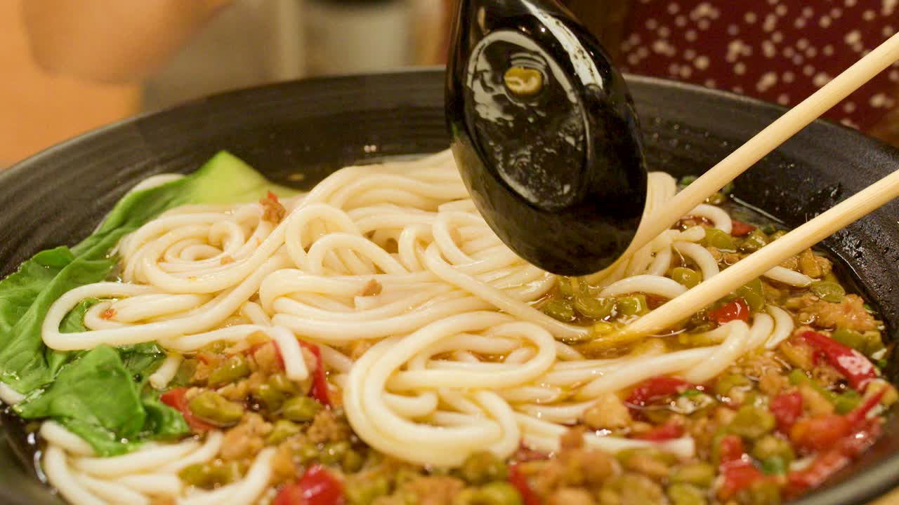 Hand stirs hot spicy noodle soup with chopsticks and spoon in bright restaurant lighting