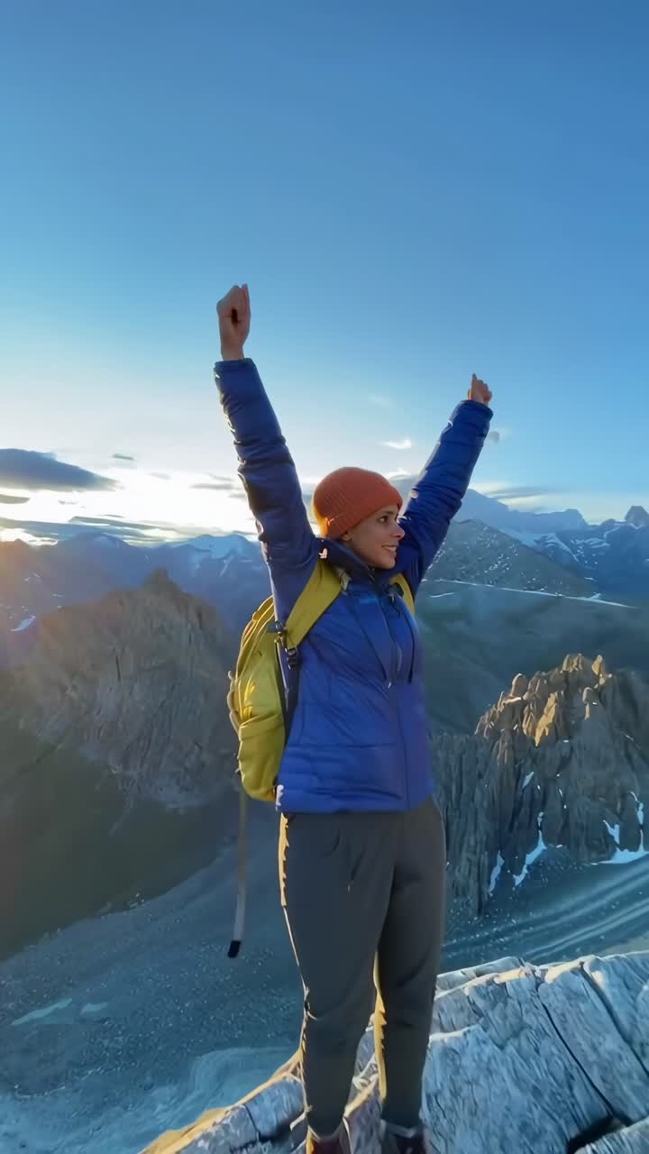Video aereo di un escursionista con giacca blu e zaino giallo su una cima rocciosa di montagna all'alba.