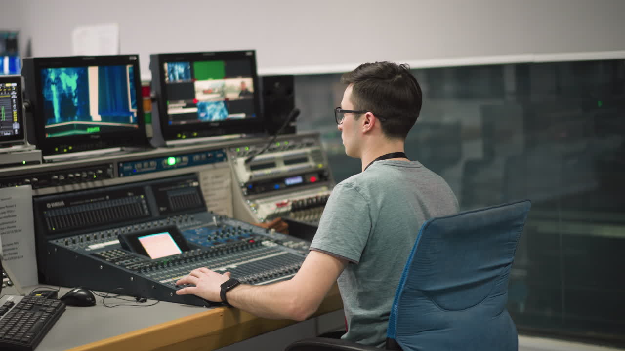 Broadcast operator controlling mixing desk and computer monitors in television studio, focusing on production equipment for media control and television broadcasting