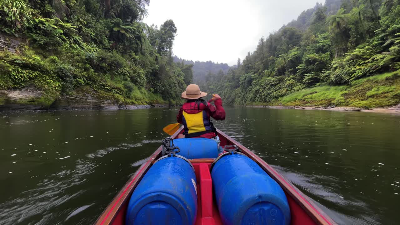 Donna con giacca rossa, cappello e giubbotto di salvataggio giallo pagaia velocemente lungo il fiume Whanganui nella sua canoa tra due scogliere boscose