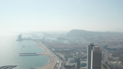 Aerial-descending-shot-of-sea-coast-with-beach-and-harbour-in-city.-Revealing-high-rise-buildings.-Hazy-view-against-sun.-Barcelona,-Spain