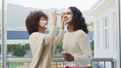 happy diverse female friends applying beauty face mask at balcony