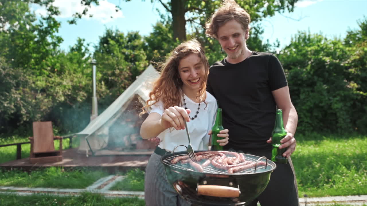Giovane coppia che frigge la carne alla griglia bevendo birra e divertendosi. Tanto verde intorno. Campeggio di lusso. Rallentatore