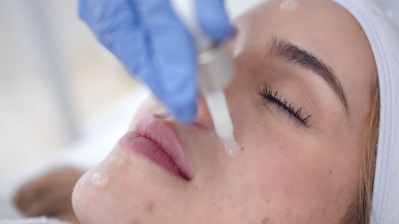 Beautiful slow motion close-up shot of a beautiful young woman's face while receiving a dermatological beauty treatment with a dropper serum inside a clinic.