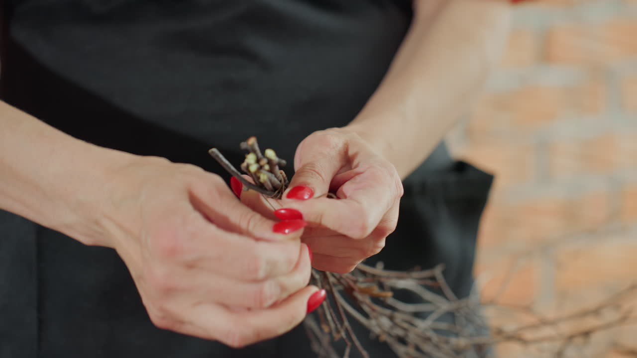 Female florist tying bundle of thin dry twigs with hands, preparing natural decoration craft detail with care, focusing on arrangement process, showcasing handmade artisan floral creativity in cozy studio