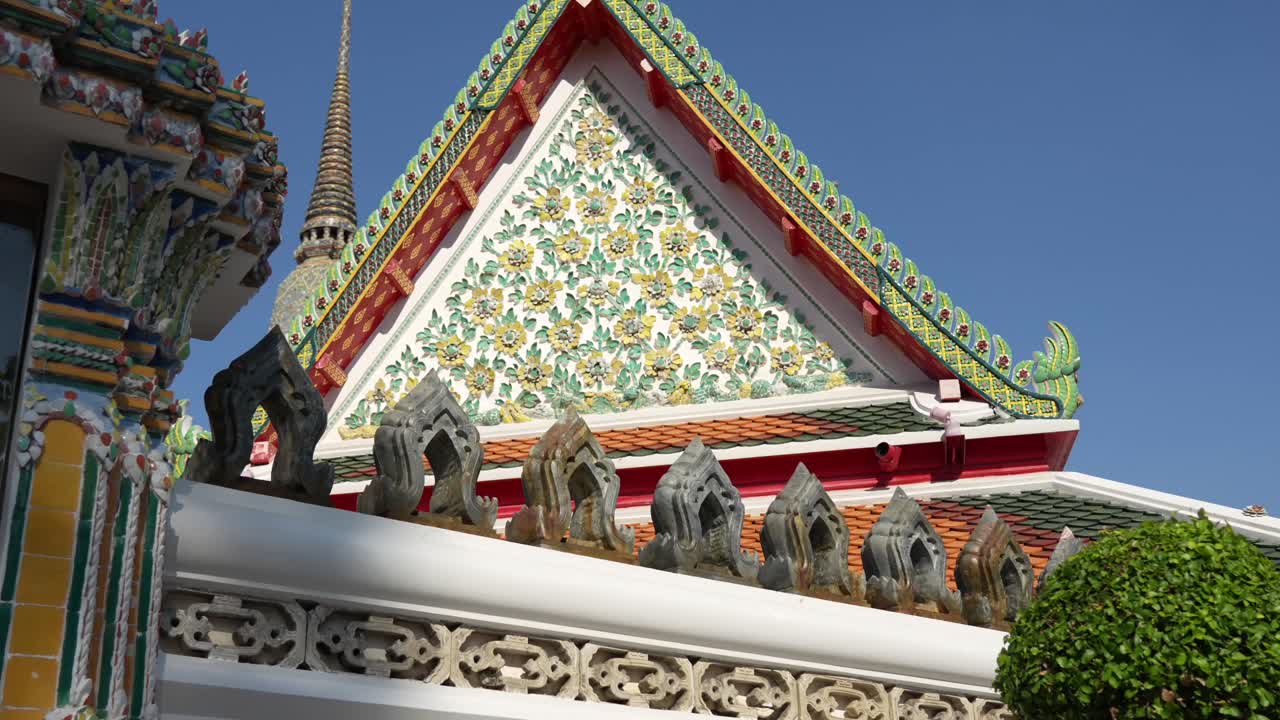 Close-up of the ornate gable end (pediment) of a Wat Pho, Bangkok structure, featuring vibrant ceramic tile mosaics of floral patterns under clear blue sky. Thai temple architecture and fine artistry