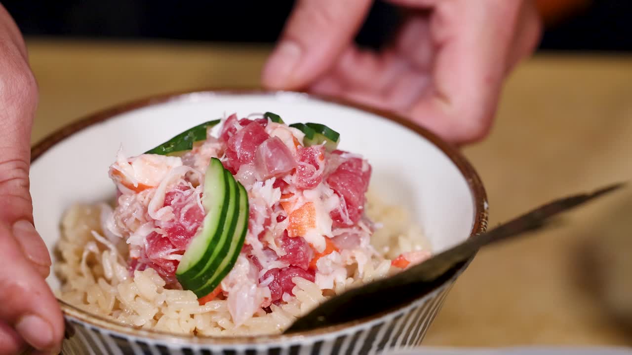 Close-up shot of a chef's hands garnishing a fresh seafood rice bowl with sliced cucumber