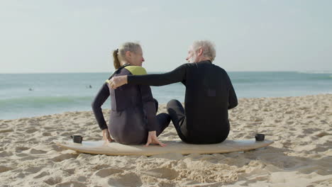 back view of a senior couple in wetsuit embracing while sitting on a surfboard at the beach