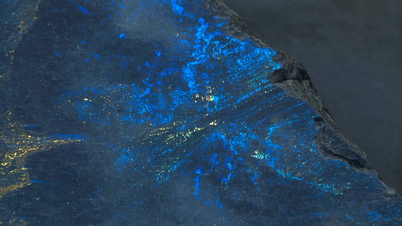 Vista macro di una lastra minerale iridescente dove la luce radente rivela riflessi blu cangianti, fratture lucide, pagliuzze d'oro e una profondità scura e texturizzata sulla superficie cristallina.