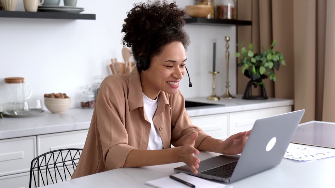 Positive successful african american curly-haired girl, in casual clothes, working or studying from home, using laptop and headset, talking on video conference, greetings, smiling friendly