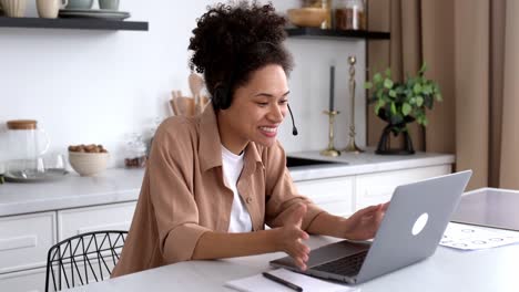 exitosa chica afroamericana de cabello rizado, con ropa casual, trabajando o estudiando desde casa, usando portátil y auriculares, hablando en videoconferencia, saludos, sonriendo amistosamente