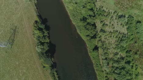 Aerial-view-over-a-river,-surrounded-by-trees-and-parkland,-with-electricity-wires-from-a-transmission-tower-crossing-the-water