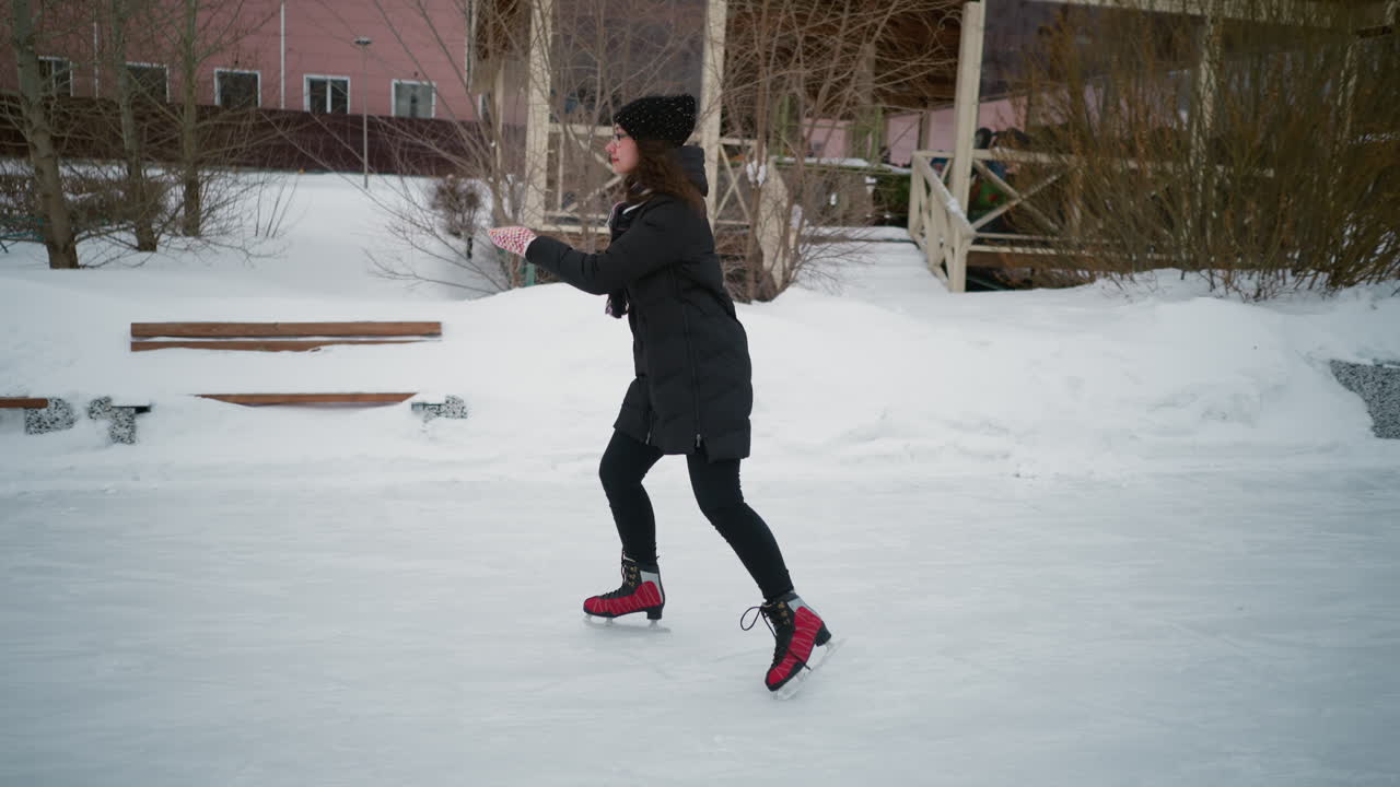 Lady skating gracefully on outdoor ice rink in winter, wearing black coat, knit hat, striped scarf, red skates, moving confidently on frozen surface surrounded by snowy landscape and rustic background