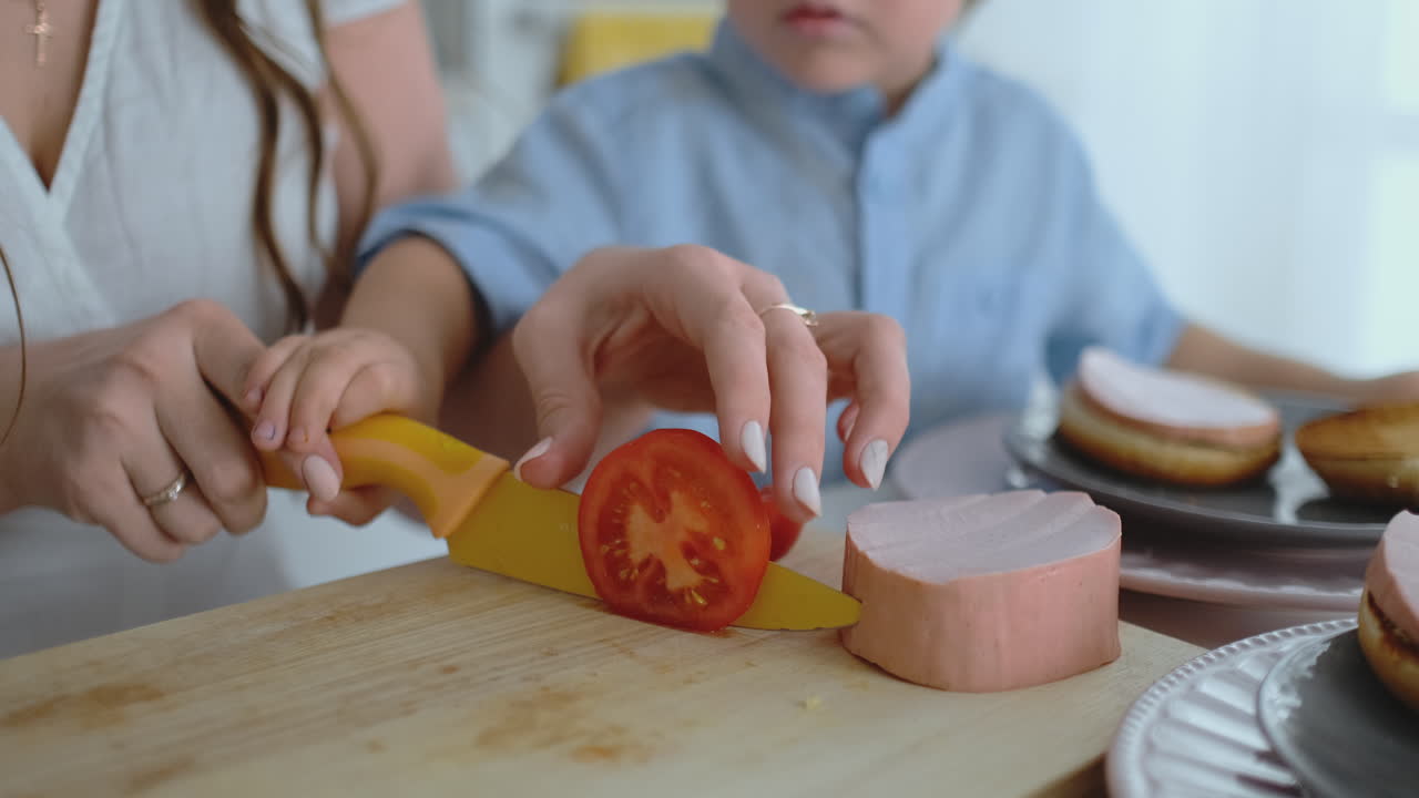 어린 아이와 함께 어린 어머니가 집에서 만든 햄버거를 위해 토마토를 칼로 잘라 함께 건강한 음식을 요리합니다.