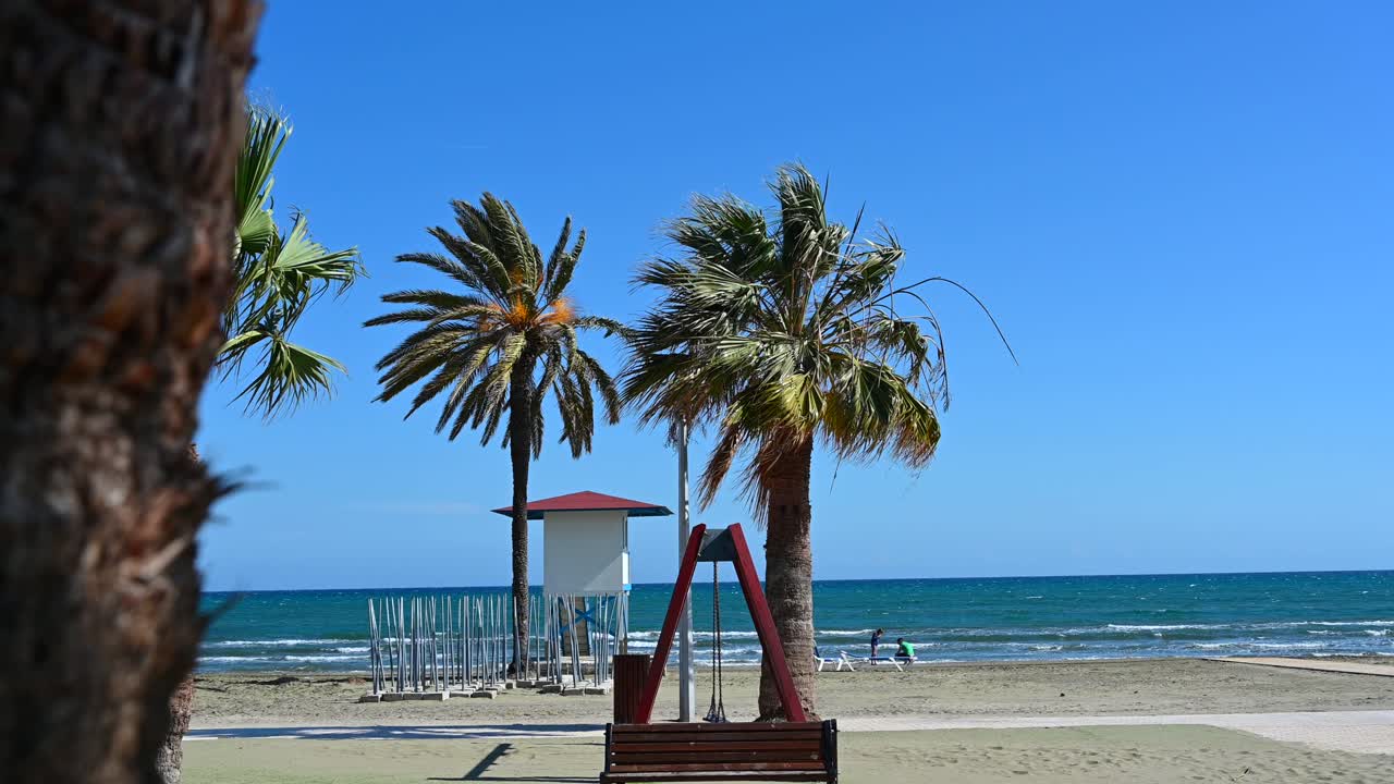 Palm trees and a white lifeguard hut with a red roof on the sandy beach of Larnaca, Cyprus, with waves rolling in and people in the distance