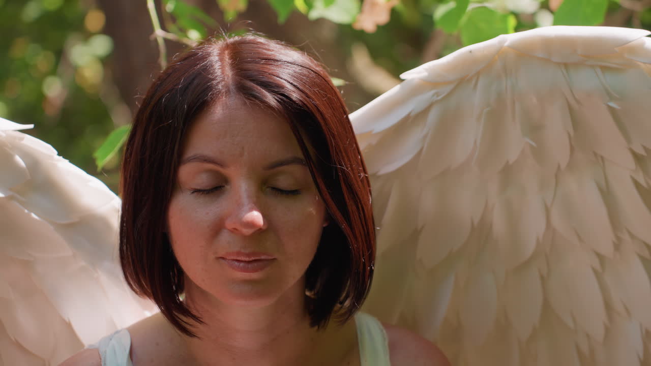 Close up upward view of fairy white goddess meditating in forest, hands resting calmly on knees, soft sunlight glowing on white dress and wings