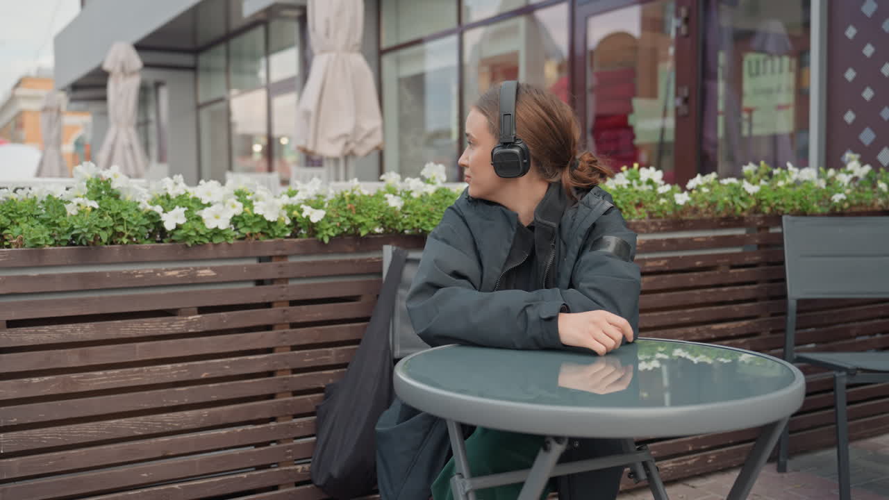 Donna con cuffie seduta al tavolino di un bar, avvolta in una giacca scura su un patio esterno accanto a una fioriera di fiori bianchi, da sola, con un sorriso rilassato, mani appoggiate sul tavolo di vetro, che guarda di lato
