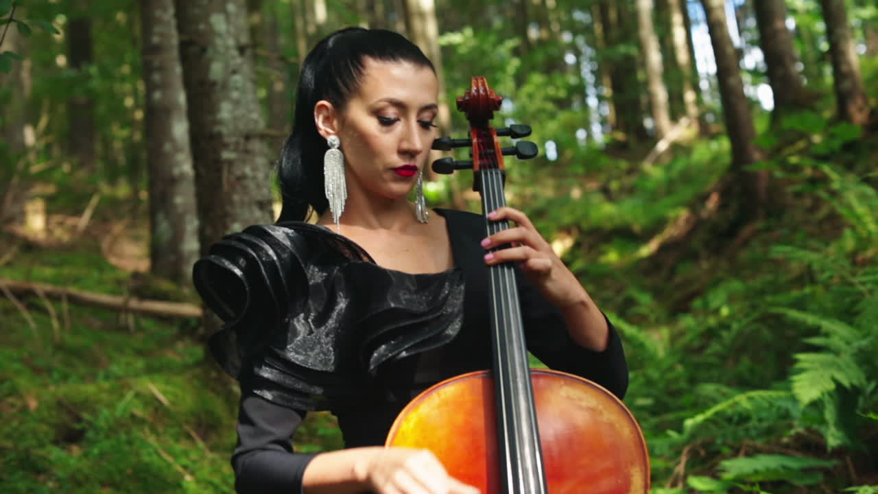 Attractive woman with musical instrument. Beautiful musician in black dress performing music in the forest. Pretty lady playing the cello outdoors.
