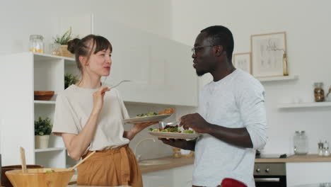 pareja multiétnica comiendo ensalada y charlando en la cocina