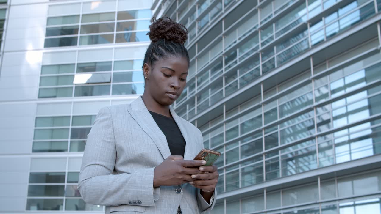 Static shot of a focused African American businesswoman standing among city office buildings and using cell phone