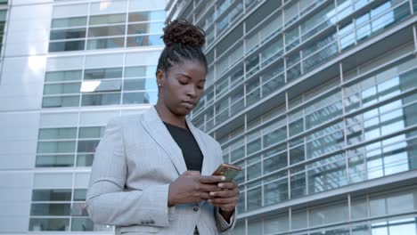 static shot of a focused african american businesswoman standing among city office buildings and using cell phone