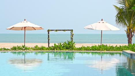 luxury tropical poolside, beach arch, parasols and reflection on pool water on sunny day, full frame