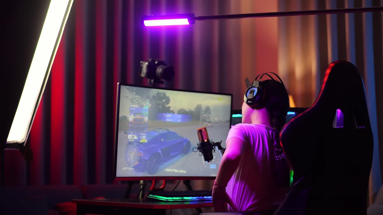 vista trasera de una chica asiática con dolor de espalda mientras juega a un juego de carreras de coches a través de la red en una computadora personal. videojuego de transmisión en vivo, escritorio iluminado por luz de tira de led rgb