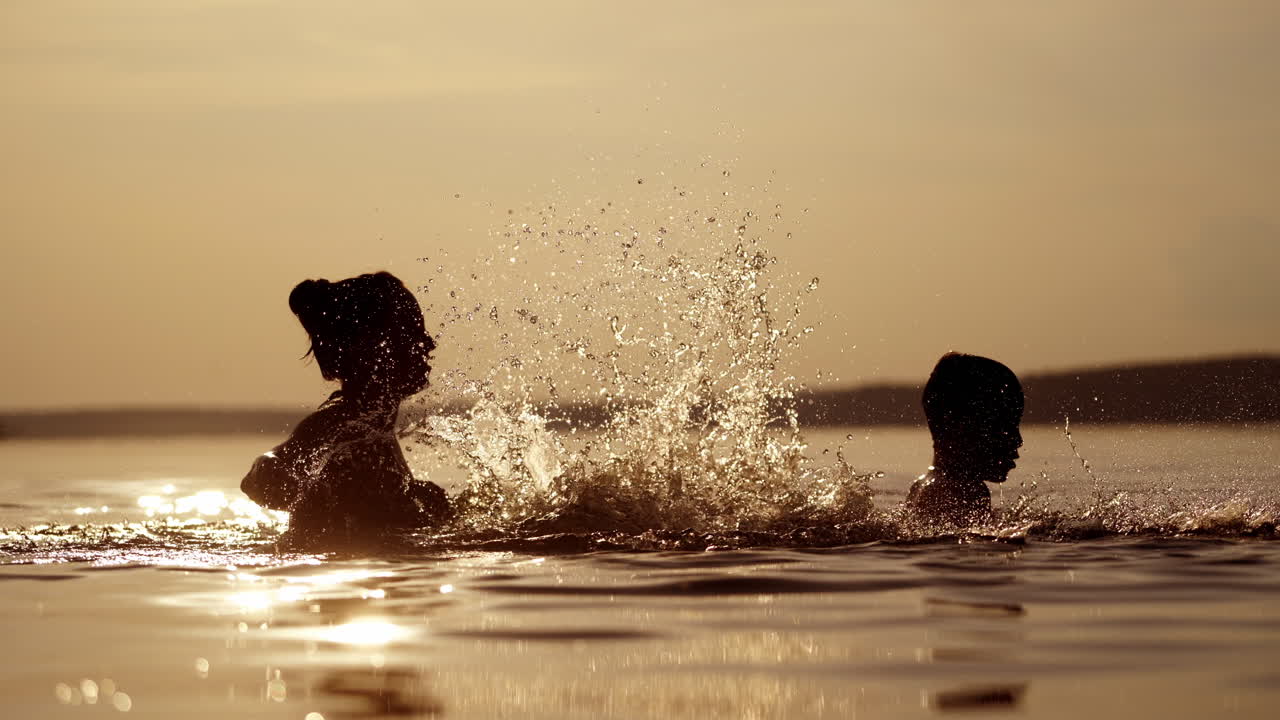 Mother and son in the river having great time together in the evening. Woman splashing water on a boy in the lake at dusk in summer time.