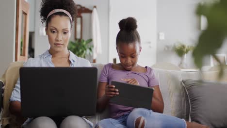 madre y hija afroamericanas felices sentadas en el sofá, usando computadora portátil y tableta, cámara lenta