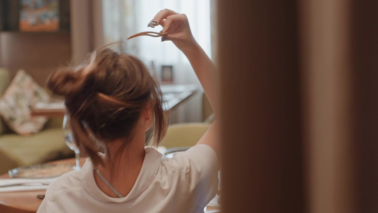 カフェでテーブルで待ちながら乱れたお団子ヘアを直す女性。グラスの反射とカーテン越しの光が差し込むビストロの雰囲気の中、まとめ髪と下ろした髪の間で切り替え、物思いにふける一時とカジュアルな雰囲気が漂う。