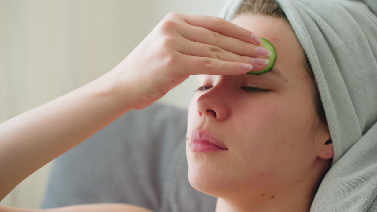 Close-up of woman rubbing cucumber on her forehead as part of a soothing skincare routine, hair wrapped in towel, creating a peaceful self-care atmosphere with natural light