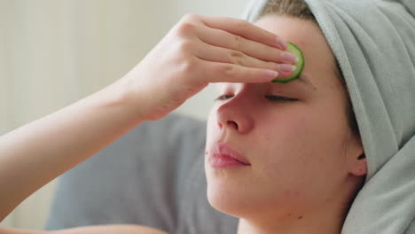 close-up of woman rubbing cucumber on her forehead as part of a soothing skincare routine, hair wrapped in towel, creating a peaceful self-care atmosphere with natural light