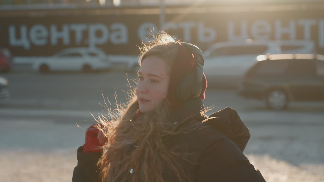 Piazza cittadina ghiacciata, giovane donna che balla con guanti rossi e paraorecchie, gesti espressivi delle mani verso il tramonto, cappotto invernale e respiro fumante, auto e sfondo della piazza, artista di strada contemplativa.