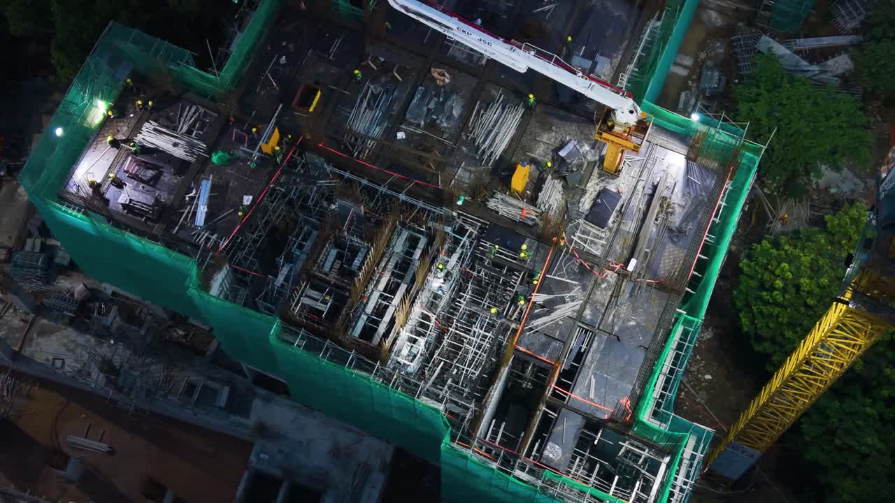 Top-down aerial of the VinHomes high-rise construction site near Hanoi's West Lake at night. Bright work lights illuminate workers and concrete pumping activity. Development and labor
