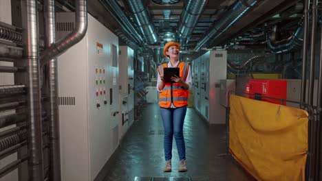 Full Body Of Asian Female Engineer With Safety Helmet Looking At The Tablet In Her Hand And Looking Around While Standing In Engine Control Room, Work Of Electrical Generators