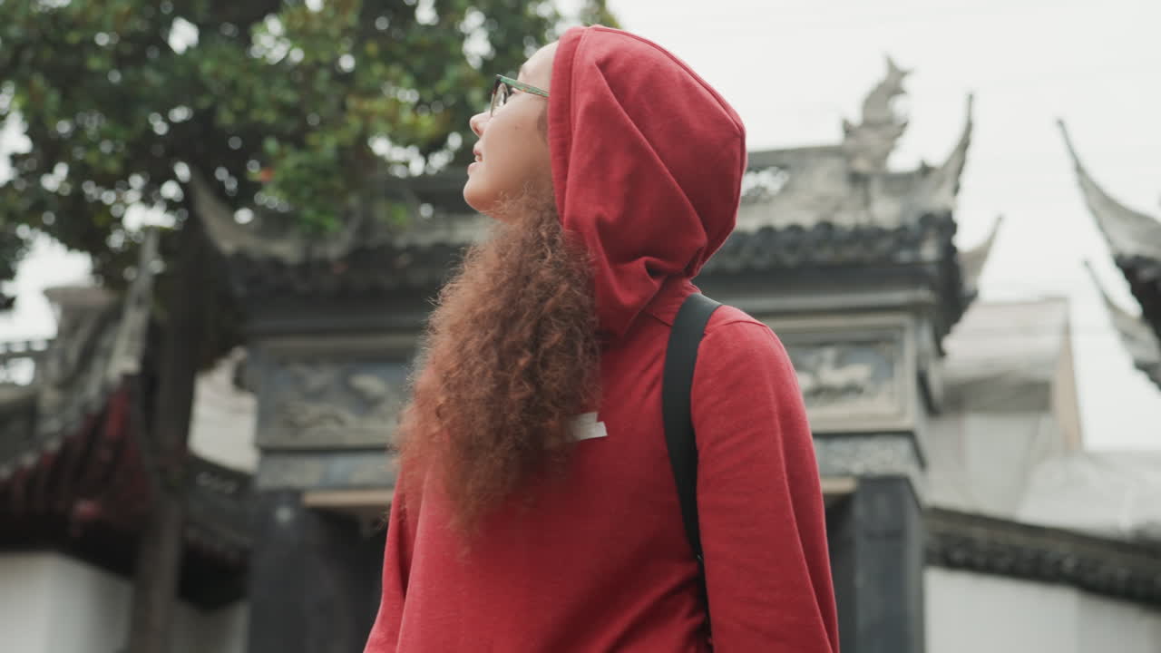 Donna di colore con felpa rossa vicino a un arco, che osserva le sagome dei tetti e i bassorilievi scolpiti, con i capelli ricci visibili sotto il cappuccio, lo zaino pronto, mentre passeggia in un cortile storico sotto un cielo coperto.