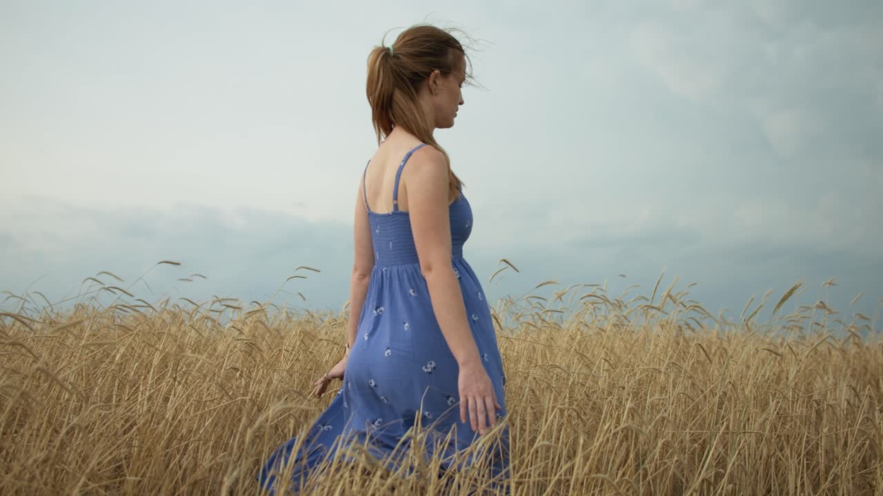 A young woman wearing a blue dress walking in a wheat field with distant storm clouds.
