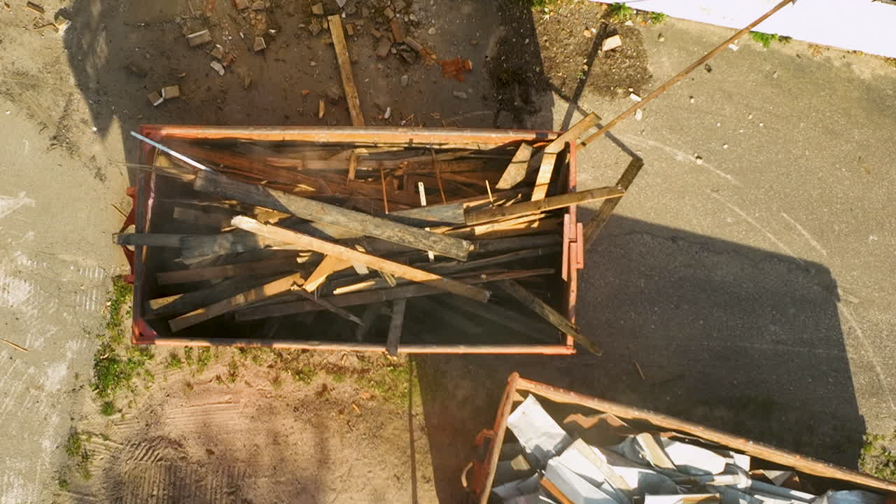 Overhead view showing an excavator loading wooden debris into a dumpster during the cleanup of a demolition site