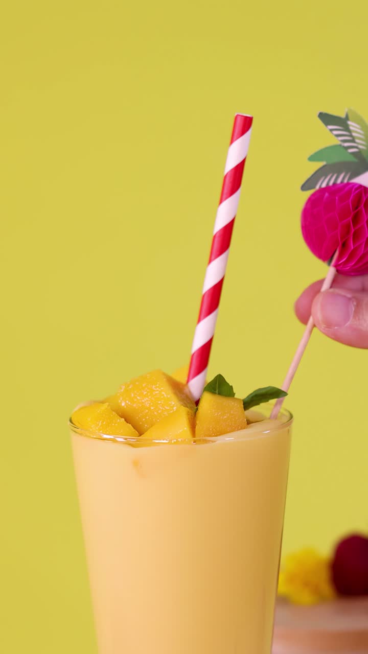 Primo piano verticale di frullato al mango mentre una mano aggiunge una cannuccia a righe e una vivace guarnizione tropicale