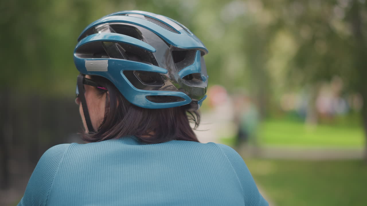 Donna vista da dietro con casco blu che percorre un sentiero del parco, maglione di lana, capelli intrecciati, luce solare soffusa che filtra tra gli alberi, profondità di campo ridotta, tranquillo tragitto urbano, stato d'animo riflessivo, spontanea