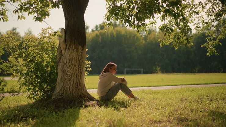 Woman sitting alone by a tree in a sunlit park, with serene expression and sunlight casting gentle shadows around her, background shows lush greenery, trees, and distant football goalpost