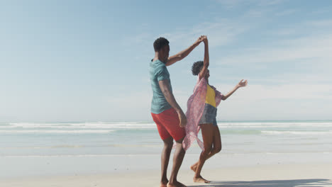 feliz pareja afroamericana bailando y tomándose de las manos en una playa soleada