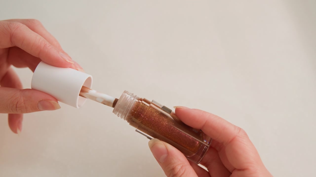 Due mani rimuovono con cura il tappo da una boccetta di lucidalabbra, rivelando un lucidalabbra metallico marrone-bronzo che ricopre l'applicatore e il liquido visibile all'interno del contenitore di vetro trasparente, mentre si avvita e si tira.