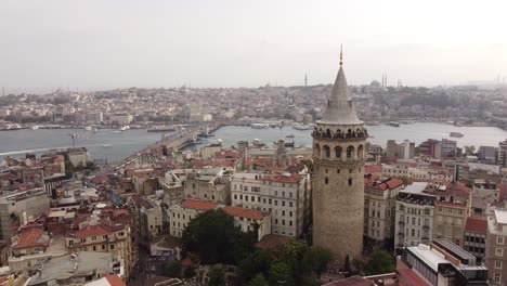 Vista-Aérea-De-La-Histórica-Torre-De-Gálata-En-Beyoglu-Estambul-Con-El-Mar-Del-Bósforo,-Mezquitas,-Tejados-Y-El-Puente-De-Gálata-Al-Fondo-Detrás