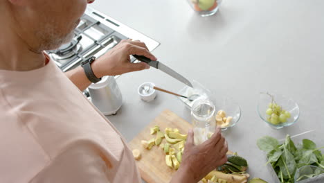 High-angle-of-senior-biracial-man-preparing-fruit-and-vegetable-smoothie-in-kitchen,-slow-motion