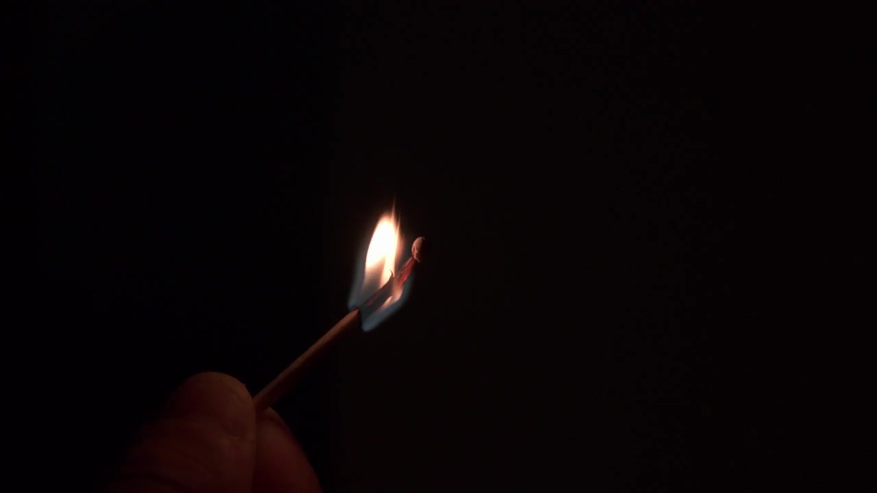 Hand holds a single match as the flame flares to life and burns against a dark background in close up.