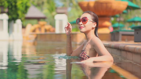 Pretty-Asian-model-girl-adjusting-sunglasses-inside-swimming-pool-on-blurred-background-shallow-focus-handheld-in-slow-motion